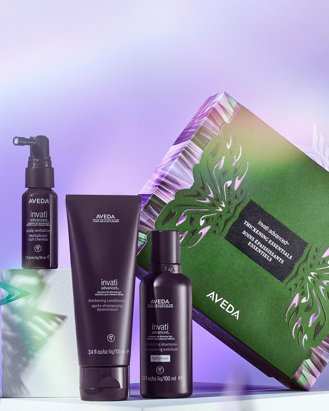 Aveda-Haarpflegeprodukte vor hellem Hintergrund. Das Set enthält ein Kopfhaut-Revitalisierendes Spray, eine verdickende Spülung und ein Peeling-Shampoo. Die Verpackung besteht aus schwarzen Flaschen, rechts befindet sich eine grün-violette Geschenkbox mit Blattmotiven.